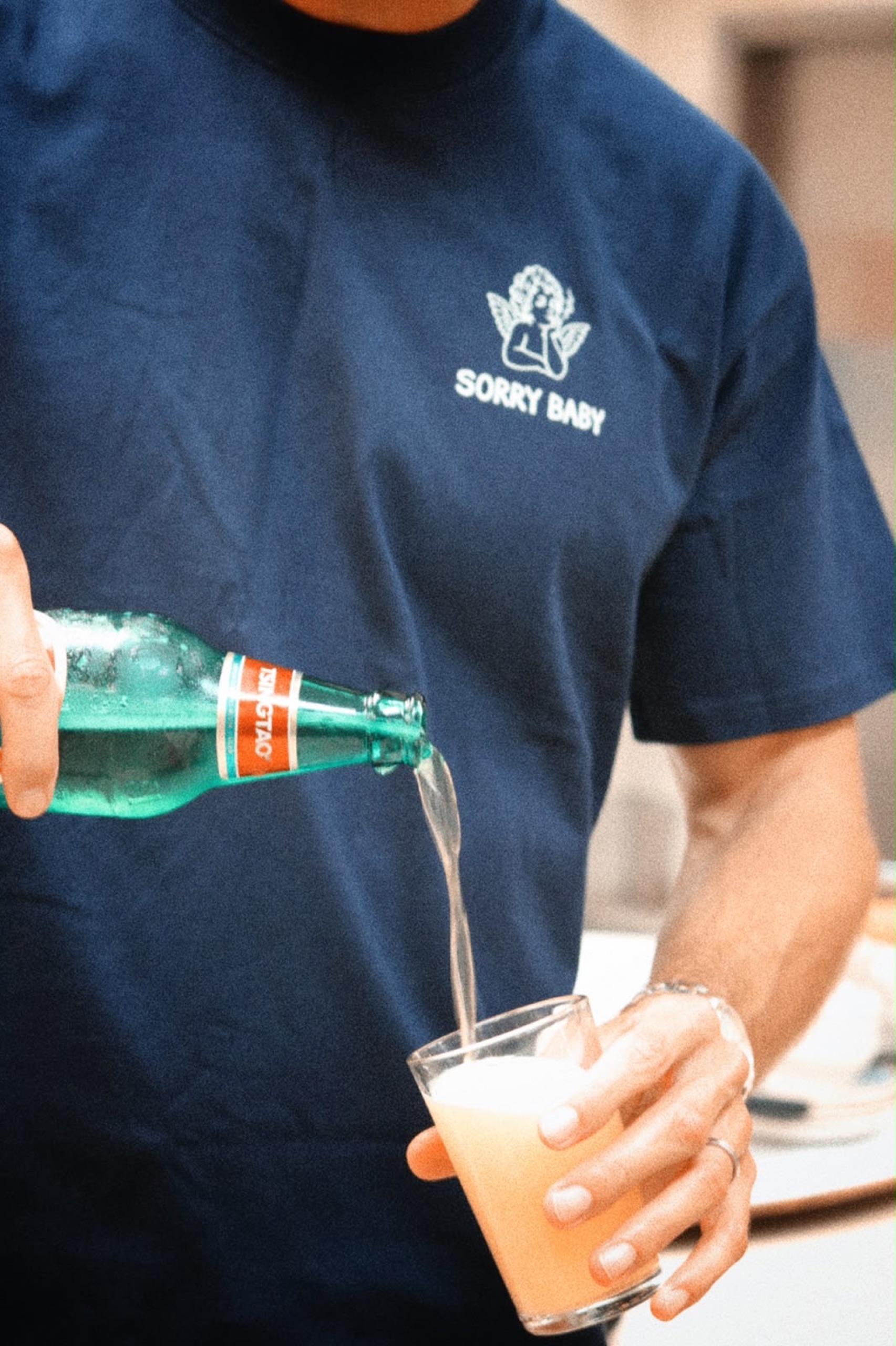 Navy SORRY BABY shirt front logo worn while pouring a Tsingtao beer