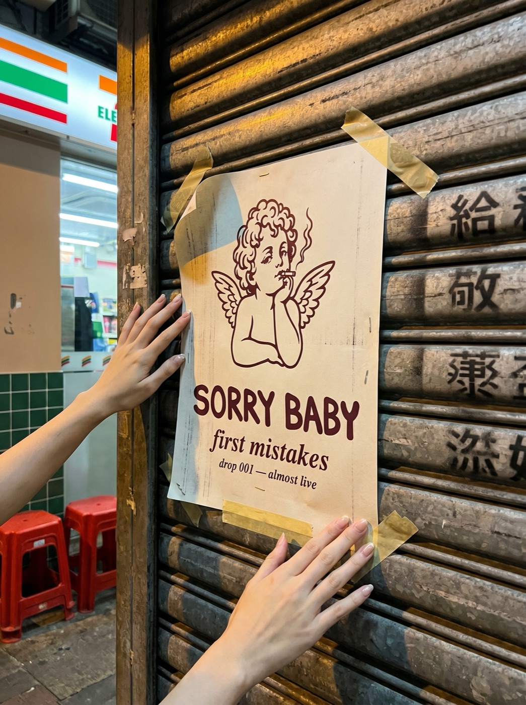 Hand-made SORRY BABY drop 001 poster taped to a Hong Kong metal shutter at night