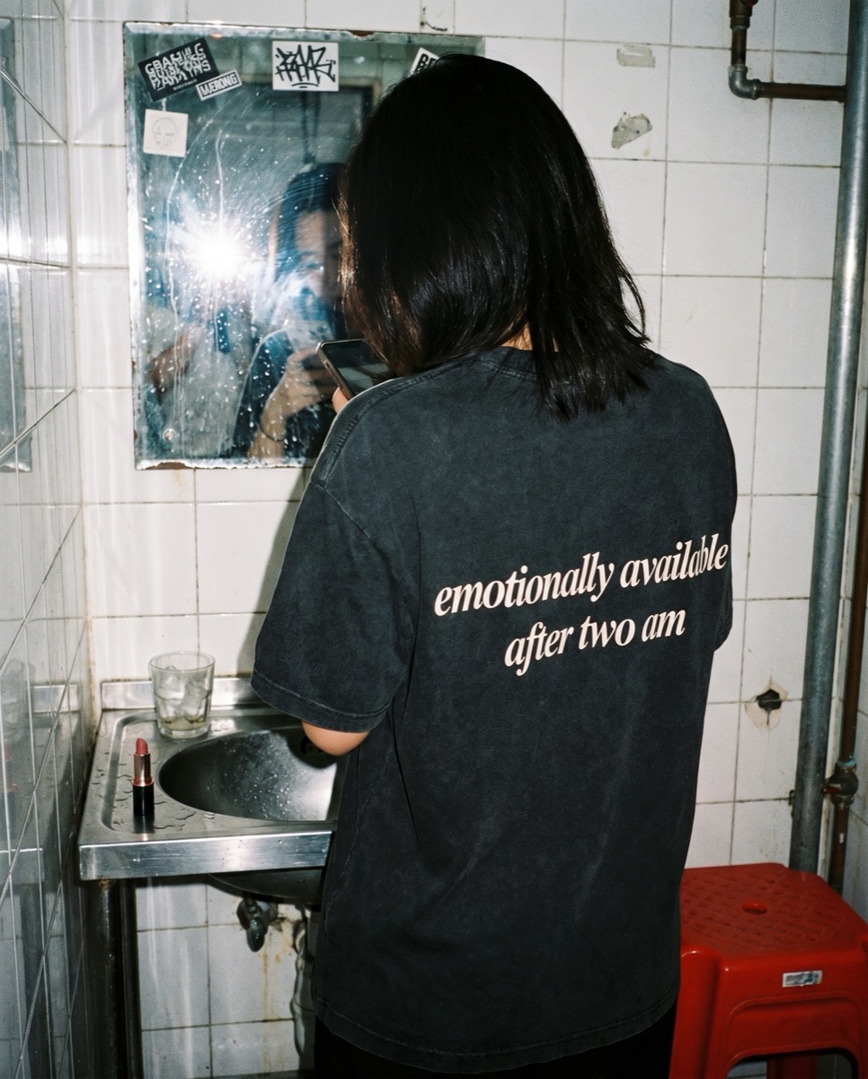 Washed black SORRY BABY shirt reading emotionally available after two am in a Hong Kong bar bathroom mirror