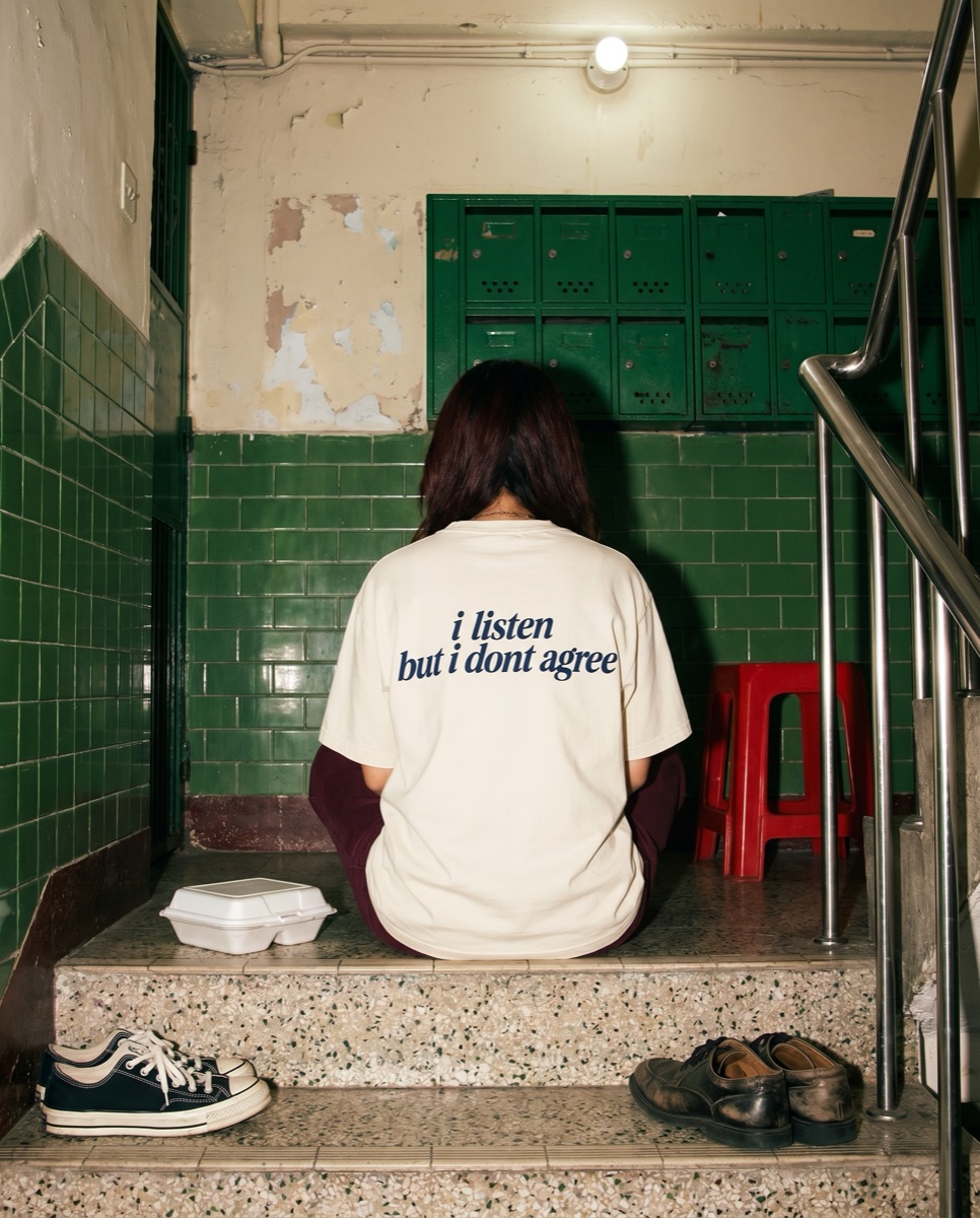 Cream SORRY BABY shirt reading i listen but i dont agree on the stairs of a Hong Kong walk-up with green tiles