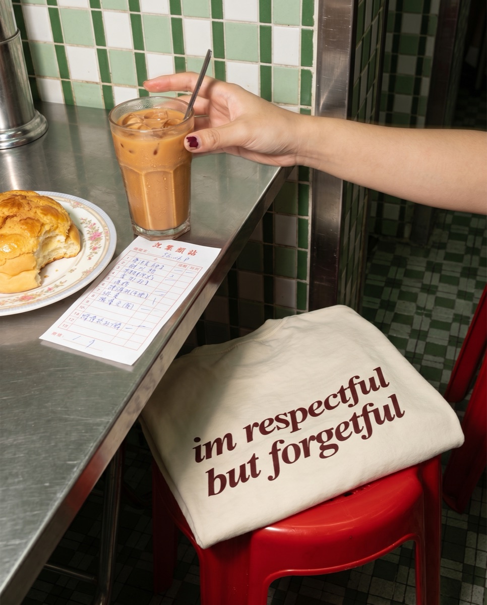 Cream SORRY BABY shirt reading im respectful but forgetful folded over a red stool in a Hong Kong cha chaan teng